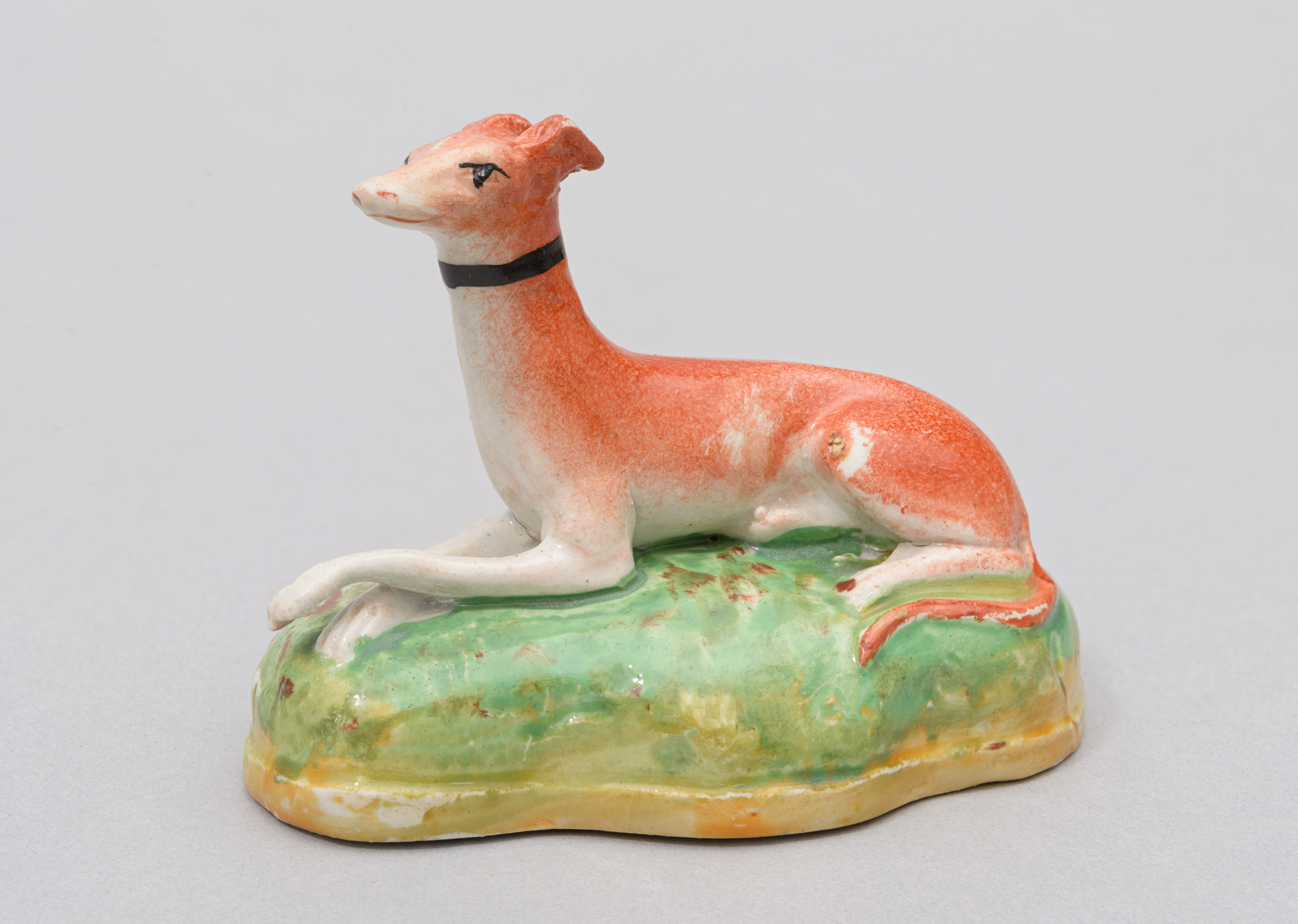 A ceramic figure of a greyhound dog with red coat and a black collar. The greyhound is sitting on a green grassy patch.